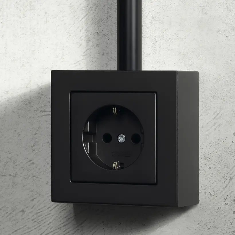 JUNG A CUBE socket in matt graphite on a light concrete wall with a black conduit