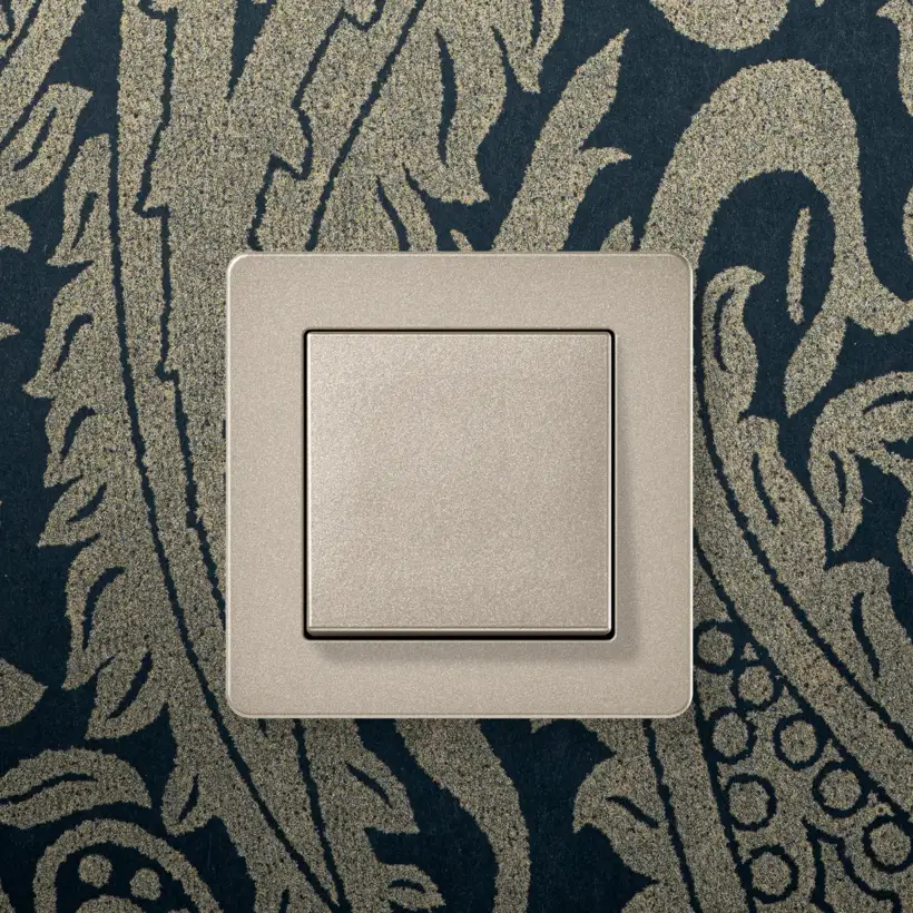 JUNG A FLOW push-button in champagne on a patterned wallpaper